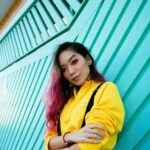 Fashionable young woman with pink hair in a yellow outfit posing against a teal wall outdoors.