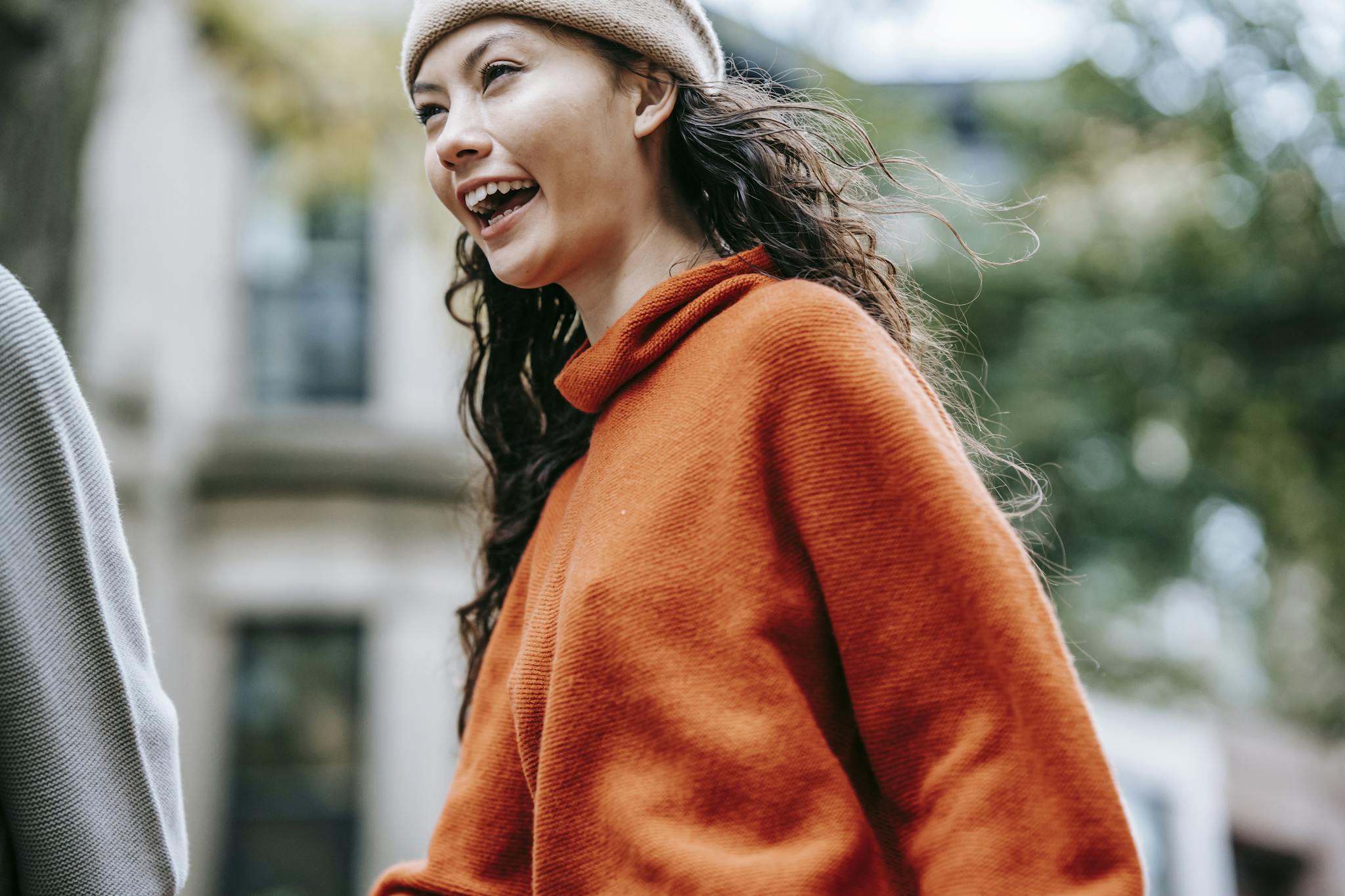 From below of laughing young Hispanic woman wearing warm hat and red sweater walking with friend along city street