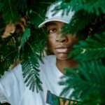 Pensive young African American male millennial in t shirt and cap standing among green plants and looking at camera